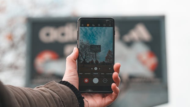 A hand holds a smartphone taking an autumn photo with trees in the background.