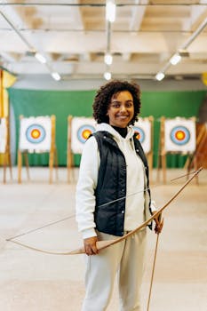Smiling young woman holding a bow in an indoor archery range with colorful targets.