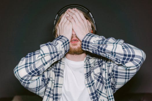 Young man wearing headphones and plaid shirt covering eyes, expressing stress or fatigue indoors.